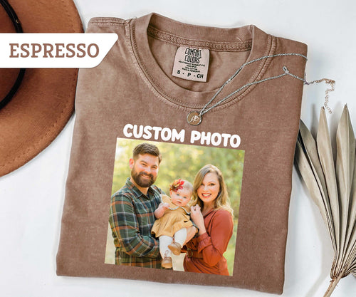 May include: An espresso-colored short-sleeve t-shirt featuring a printed custom photo of a family of three. The shirt has a Comfort Colors brand label at the inner neckline and the text "CUSTOM PHOTO" printed 