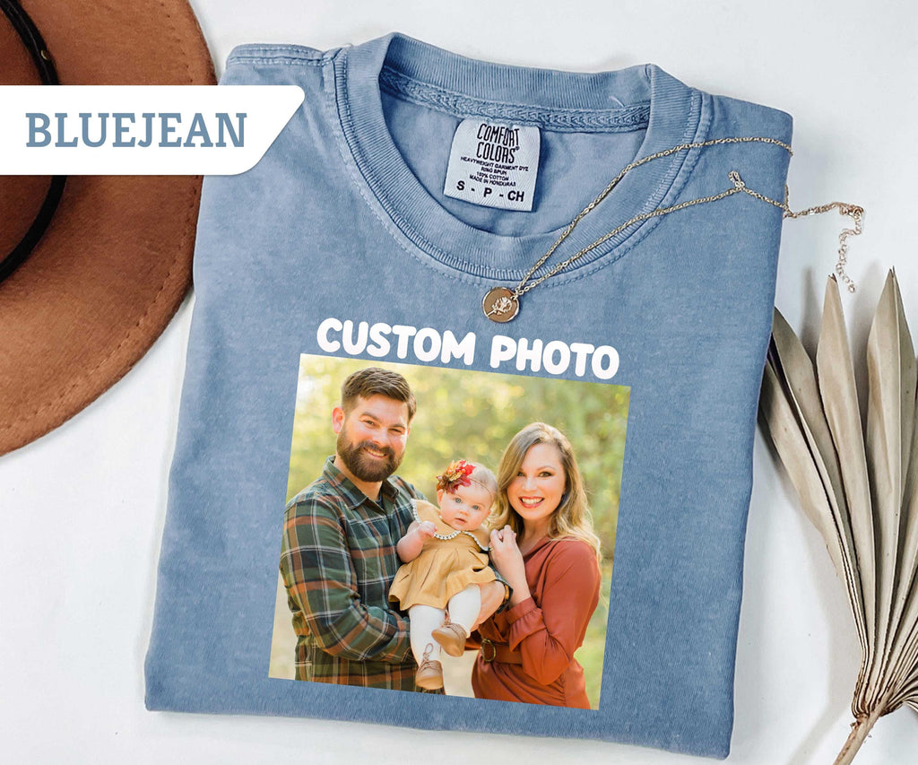 May include: A blue jean colored short-sleeve t-shirt featuring a printed rectangular photograph of a family consisting of a man, a woman, and a baby. The text CUSTOM PHOTO is printed in white above the image. The shirt 