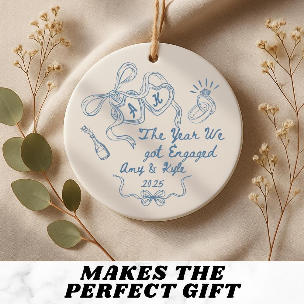 May include: A round, white ceramic ornament with a blue design and a jute string for hanging. The ornament features a drawing of a ring, champagne bottle, and ribbons. Text on the ornament reads "The Year We got En