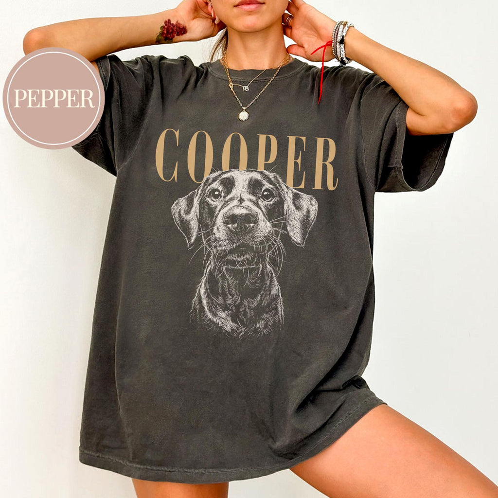 May include: A charcoal gray short-sleeve t-shirt featuring a graphic of a dog's face in the center. Above the dog, the text COOPER is printed in a tan serif font. A circular badge in the top left corner contains th