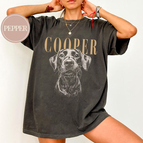 May include: A charcoal gray short-sleeve t-shirt featuring a graphic of a dog's face in the center. Above the dog, the text COOPER is printed in a tan serif font. A circular badge in the top left corner contains th
