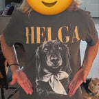 May include: A charcoal gray short-sleeve t-shirt featuring the text HELGA in large gold letters above a detailed black and white graphic illustration of a long-haired dachshund wearing a bow tied around its neck.