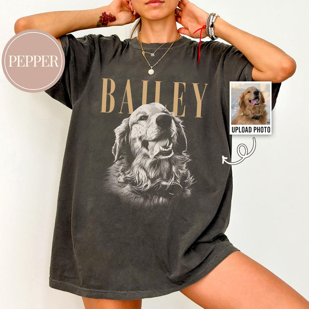 May include: A charcoal gray short-sleeve t-shirt featuring a large black and white graphic of a golden retriever dog. The name BAILEY is printed in tan capital letters above the dog. A small circular tag reads PEPPER.