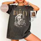May include: A charcoal gray short-sleeve t-shirt featuring a large black and white graphic of a golden retriever dog. The name BAILEY is printed in tan capital letters above the dog. A small circular tag reads PEPPER.