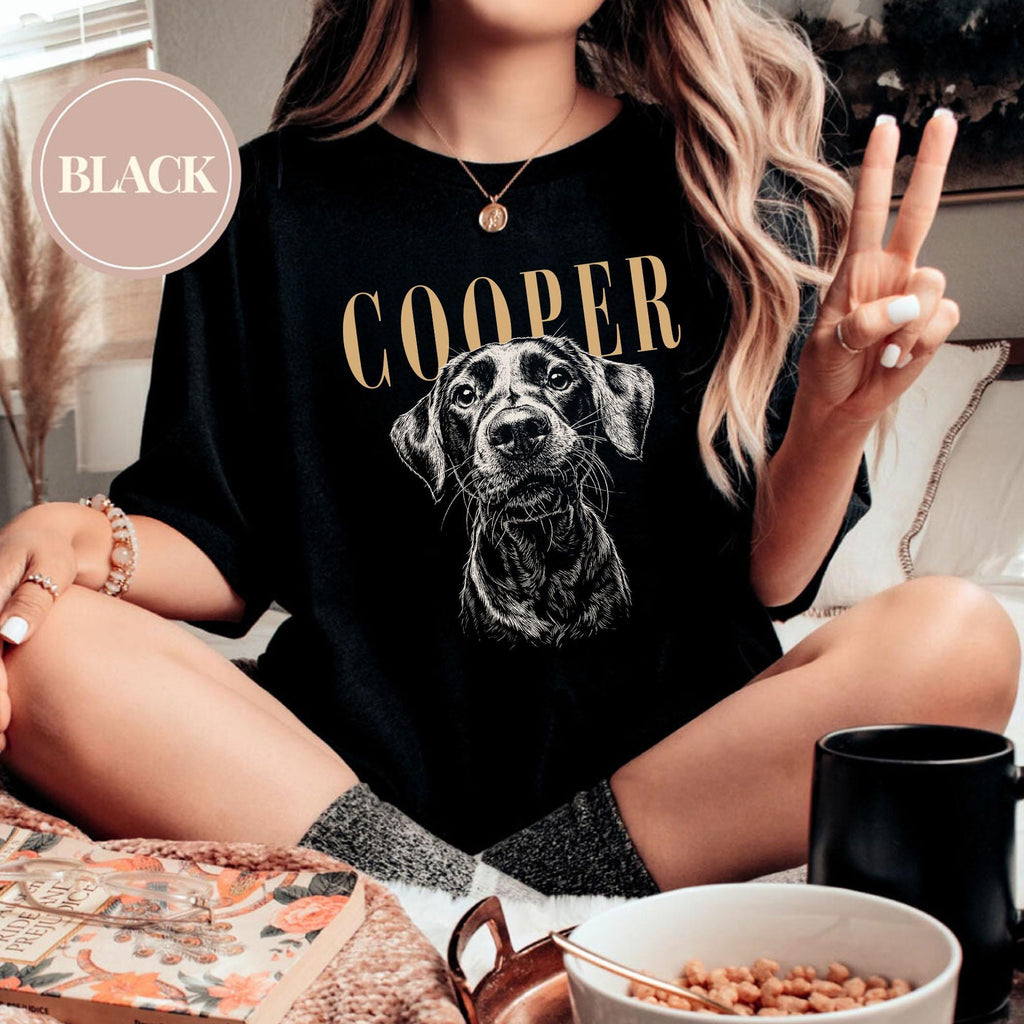 May include: A black t-shirt featuring a white line-art illustration of a dog's face centered below the text COOPER printed in gold. The shirt has a crew neckline and short sleeves. A circular badge in the top left 