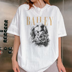 May include: A white short-sleeve t-shirt featuring a black and white illustration of a golden retriever dog with its tongue out. Above the dog, the name BAILEY is printed in large, gold-colored capital letters. The shir