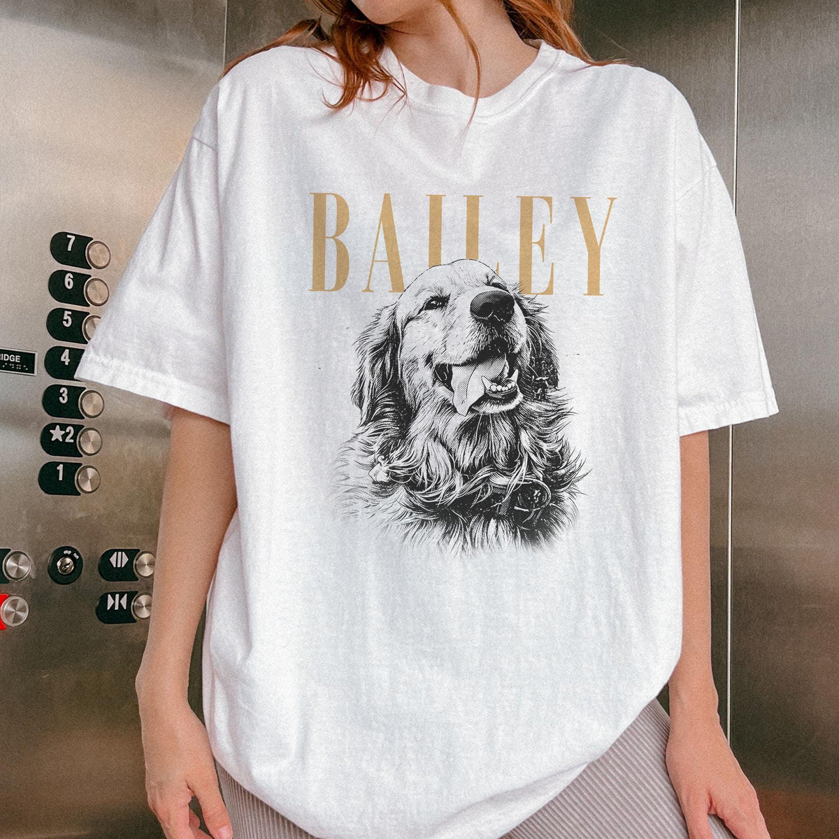 May include: A white short-sleeve t-shirt featuring a black and white illustration of a golden retriever dog with its tongue out. Above the dog, the name BAILEY is printed in large, gold-colored capital letters. The shir