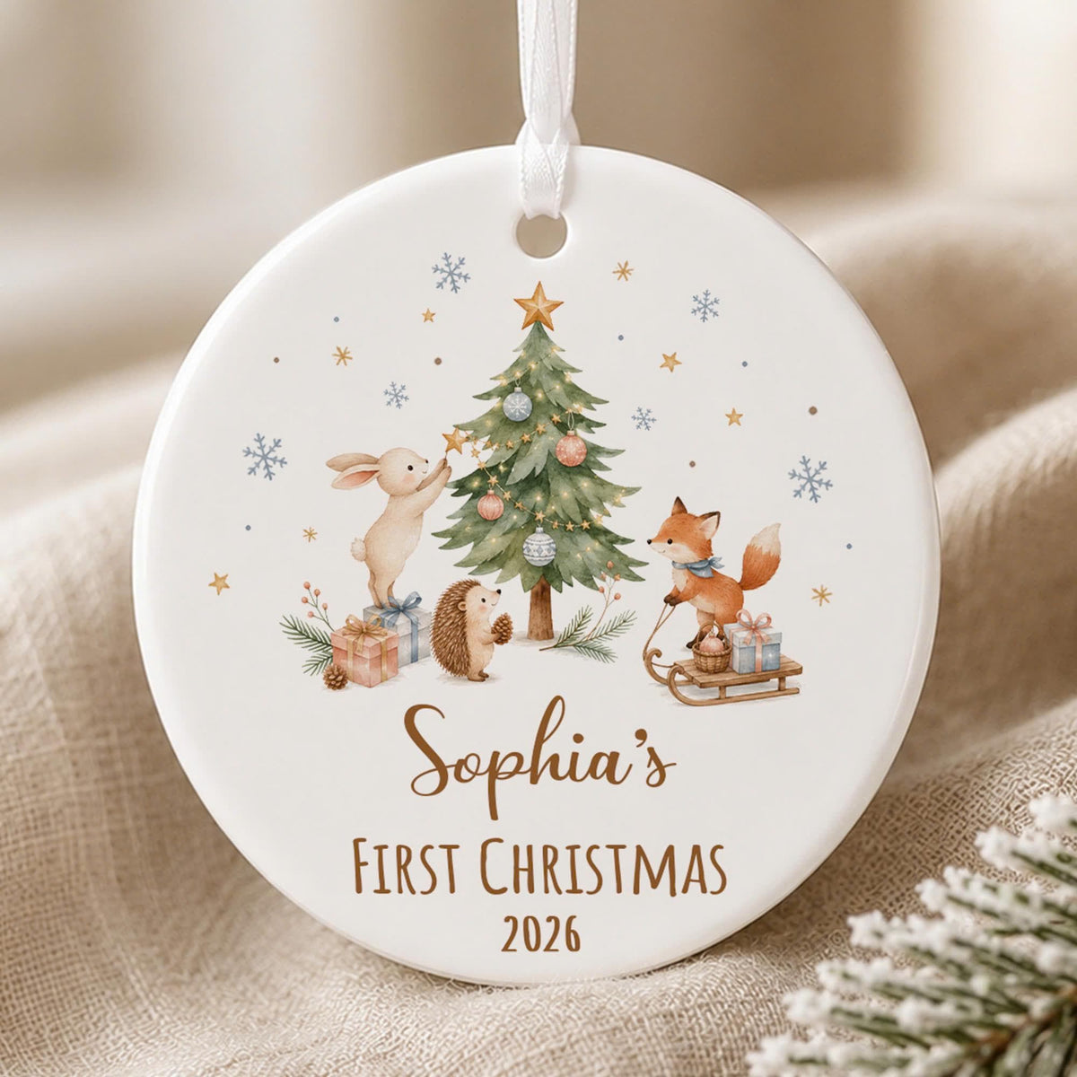 May include: A round white ceramic ornament with a white ribbon hanger featuring a watercolor illustration of a rabbit, hedgehog, and fox decorating a Christmas tree with gifts, snowflakes, and the text Sophia's Fir