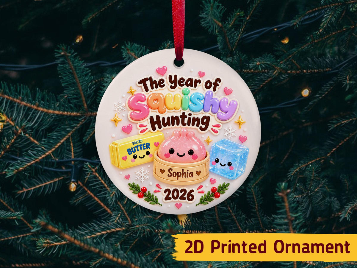 May include: A round white ceramic ornament with a red ribbon featuring the text The Year of Squishy Hunting, Sophia, and 2026. The design includes a yellow butter stick, a pink dumpling in a steamer, and a blue ice cube