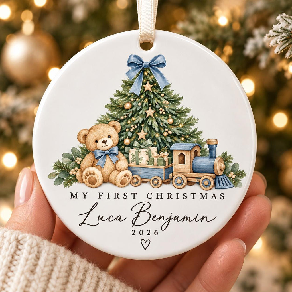 May include: A round white ceramic ornament featuring a brown teddy bear, a green Christmas tree, and a blue toy train. The text reads "MY FIRST CHRISTMAS", "Luca Benjamin", and "2026" with 