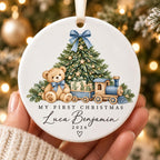 May include: A round white ceramic ornament featuring a brown teddy bear, a green Christmas tree, and a blue toy train. The text reads "MY FIRST CHRISTMAS", "Luca Benjamin", and "2026" with 