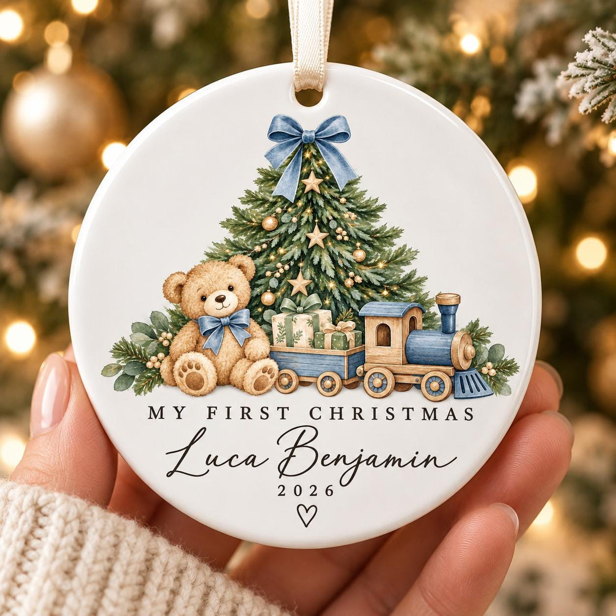 May include: A round white ceramic ornament featuring a brown teddy bear, a green Christmas tree, and a blue toy train. The text reads "MY FIRST CHRISTMAS", "Luca Benjamin", and "2026" with 