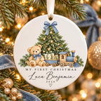 May include: A round white ceramic Christmas ornament featuring a teddy bear with a blue bow, a decorated pine tree, and a toy train. The text reads MY FIRST CHRISTMAS, Luca Benjamin, and 2026 with a small heart icon bel