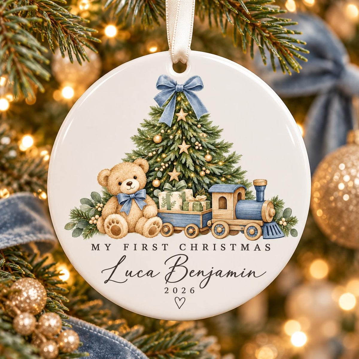May include: A round white ceramic Christmas ornament featuring a teddy bear with a blue bow, a decorated pine tree, and a toy train. The text reads MY FIRST CHRISTMAS, Luca Benjamin, and 2026 with a small heart icon bel