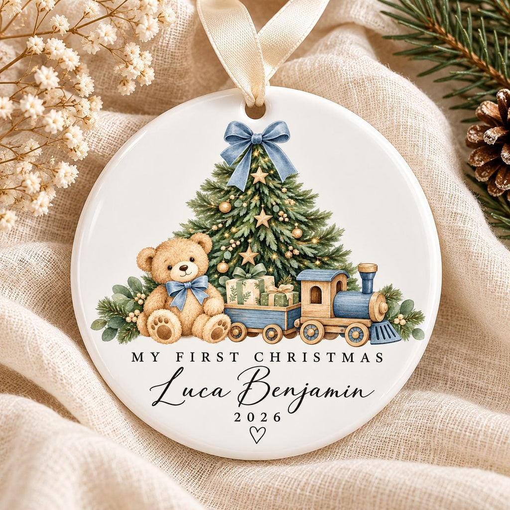 May include: A round white ceramic Christmas ornament featuring a light brown teddy bear with a blue bow, a decorated pine tree, and a blue toy train carrying gifts. The text reads MY FIRST CHRISTMAS, Luca Benjamin, and 